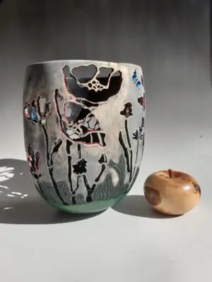 A detailed photograph of a hand-carved wooden vase named "Resilience" made from paper birch wood. The vase has an elegant, curved form with delicate cutouts of poppies and butterflies and a textured, gray painted finish. Apple shown for scale.